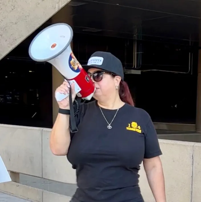 Picture of Chair, Trish Muir at an informational picket. Speaking into a white and red bullhorn and wearing a black Teamsters Local 104 t-shirt.